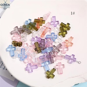 Pode incluir: Uma coleção de contas translúcidas em forma de cruz em várias cores, incluindo rosa, roxo, verde e azul. As contas estão espalhadas sobre uma superfície branca. O número "1#" é visível no canto superior direito. Adequado para fazer joias e artesanato.
