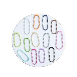May include: A collection of colorful ball chain connectors in various shades, including orange, blue, brown, and pink, arranged on a white circular surface. Other colors include green, yellow, gray, purple, black, and light blue. These connectors are used for keychains and other accessories.