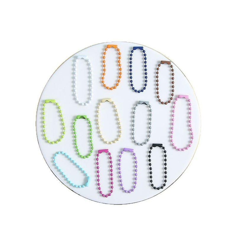 May include: A collection of colorful ball chain connectors in various shades, including orange, blue, brown, and pink, arranged on a white circular surface. Other colors include green, yellow, gray, purple, black, and light blue. These connectors are used for keychains and other accessories.