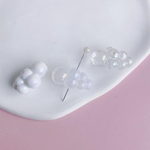 May include: Three clusters of iridescent white beads, each cluster is shaped like a cloud. The beads are on a white surface.