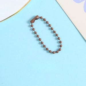 May include: A brown metal ball chain keychain with a clasp. The chain is made of small, evenly spaced, round beads. The background is a light blue surface with a hint of white and purple.