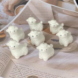 May include: Seven small, fluffy white sheep charms, each with a silver loop for hanging. The sheep have pink facial details and are displayed on a light-coloured surface. These charms are ideal for crafting or jewellery making.