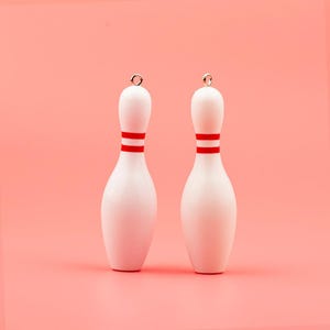 May include: Two white bowling pin ornaments with a red stripe around the middle, each with a small silver loop at the top. The pins are set against a solid pink background.