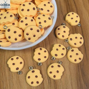 Op de afbeelding: Een verzameling gele koekjesbedels met bruine chocoladesnippers. Sommige koekjes hebben een hapje genomen. Elke bedel heeft een zilverkleurige lus voor het maken van sieraden of knutselen.