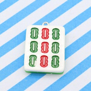May include: A white Mahjong tile pendant with green and red symbols. The pendant has a silver loop at the top for attaching to a necklace or bracelet. The tile is set against a blue and white striped background.