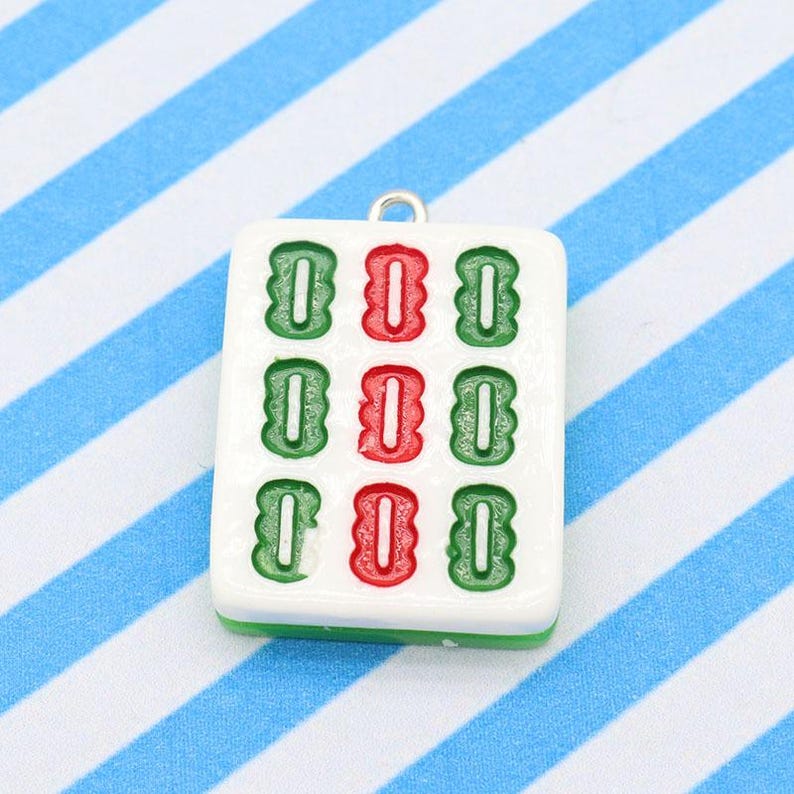 May include: A white Mahjong tile pendant with green and red symbols. The pendant has a silver loop at the top for attaching to a necklace or bracelet. The tile is set against a blue and white striped background.