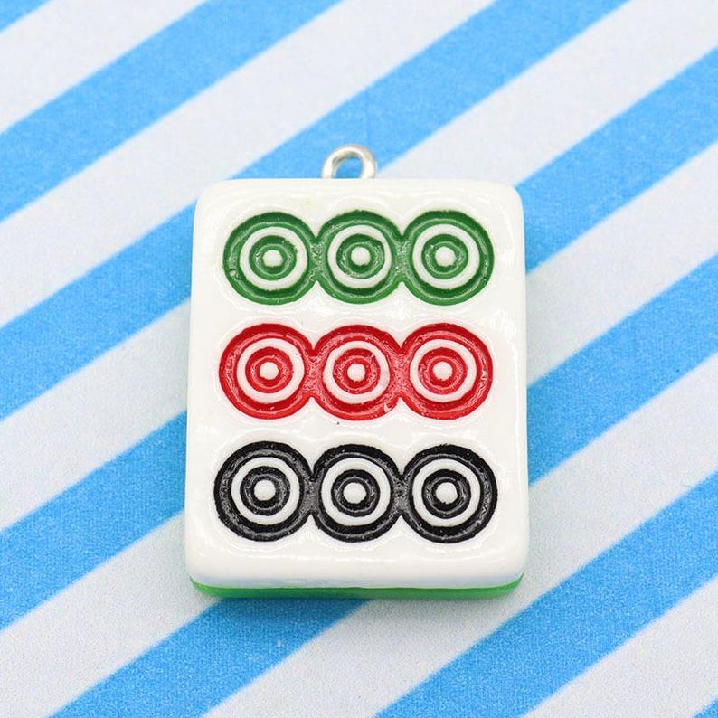 May include: A white rectangular pendant with three rows of three concentric circles in green, red, and black. The pendant has a silver loop at the top for hanging. The background is a blue and white striped pattern.