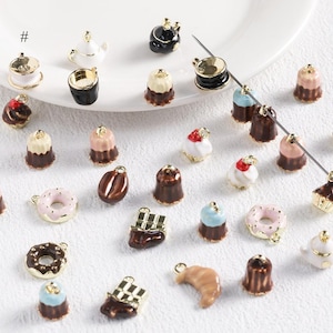 May include: A collection of miniature food charms, including donuts, cupcakes, croissants, and chocolate bars. The charms are in various colors, such as pink, brown, and gold, and are designed with intricate details. Some charms resemble teacups and teapots.