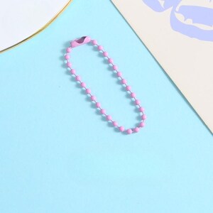 May include: A pink ball chain keychain with a clasp, lying on a light blue surface. The chain is made of small, evenly spaced pink beads. The clasp is a matching pink color. The background includes a white plate with a gold rim and a piece of paper.
