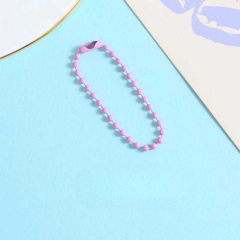 May include: A pink ball chain keychain with a clasp, lying on a light blue surface. The chain is made of small, evenly spaced pink beads. The clasp is a matching pink color. The background includes a white plate with a gold rim and a piece of paper.