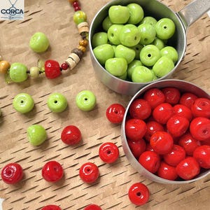 May include: Overhead shot of green and red ceramic beads, some in metal measuring cups. A partially assembled necklace with green, red, and tan beads is in the upper left corner. The beads are arranged on a brown paper surface.