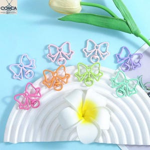 May include: A collection of colorful, openwork bow-shaped charms in shades of pink, orange, green, and purple. Each charm has a small, round pendant. The charms are arranged on a white, curved surface with a blue background and a white flower.