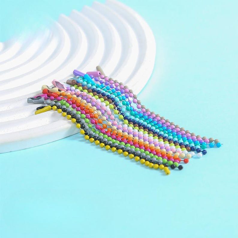 May include: A collection of colorful beaded hair clips in various shades, including yellow, black, gray, orange, pink, purple, and blue. The clips are arranged on a white, curved surface against a light blue background.