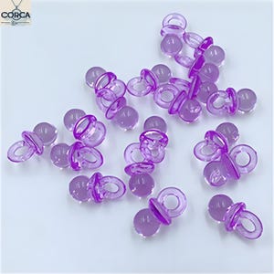 May include: A collection of small, translucent purple pacifier-shaped charms. Each charm features a round nipple, a circular shield, and a loop for attaching to a chain or other material. The charms are scattered on a white surface.