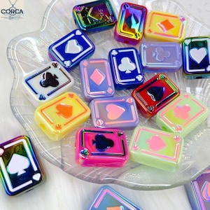 May include: A collection of colorful playing card-shaped beads. Each bead features a different suit symbol: hearts, diamonds, clubs, and spades. The beads have a glossy finish and are in various colors, including blue, pink, yellow, and green.