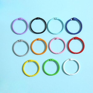 May include: A collection of colorful binder rings in various shades, including purple, black, teal, and red. The rings are arranged on a light blue background. Each ring has a small, circular opening mechanism.