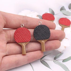 May include: Two miniature table tennis paddle charms, one red and one black, with a wood-like handle. The charms are hanging from silver loops.