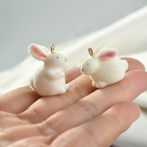 Peut inclure: Deux breloques en forme de lapin en peluche blanche avec des anneaux de saut dorés. Les lapins ont des oreilles roses et sont assis sur une main.