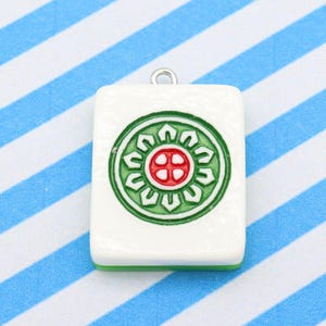 May include: A white rectangular pendant with a green and red circular design, featuring a silver loop at the top. The pendant has a green border and is set against a blue and white striped background. This pendant is likely a charm or a piece of jewelry.