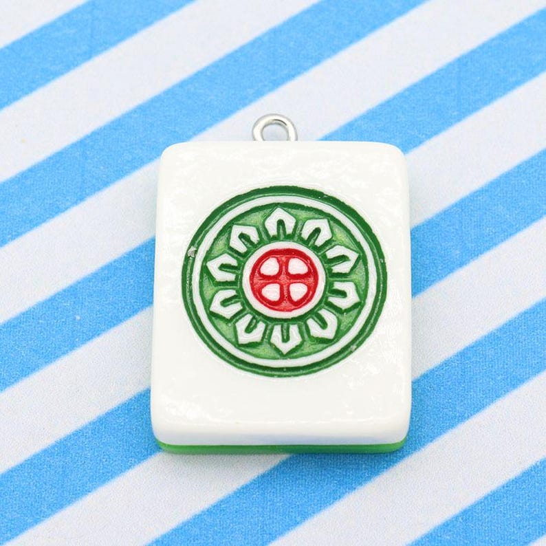 May include: A white rectangular pendant with a green and red circular design, featuring a silver loop at the top. The pendant has a green border and is set against a blue and white striped background. This pendant is likely a charm or a piece of jewelry.