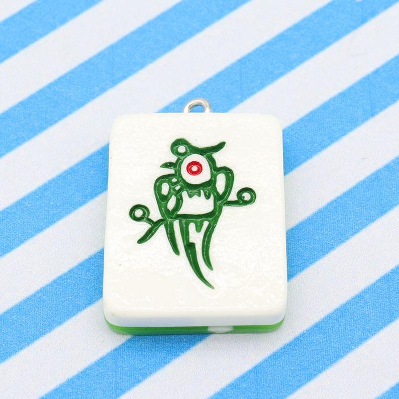 May include: A white rectangular pendant with a green and red cartoon character design. The pendant has a green border and a silver loop at the top for hanging. The background is a blue and white striped pattern.