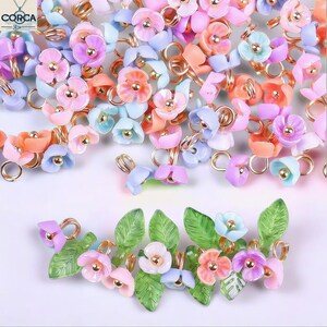 May include: A collection of colorful floral-shaped beads in pastel shades of pink, orange, blue, and purple. Each bead has a gold-colored wire loop and a small gold-colored center. Some beads are arranged with green leaf accents.