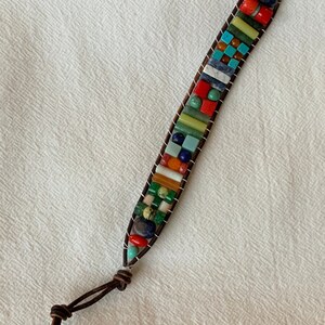 May include: A multi-colored beaded bracelet with a brown leather strap and a wooden button closure. The beads are made of various stones, including turquoise, lapis lazuli, and jasper.