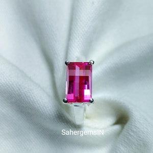 May include: A silver ring with a large, rectangular, bright pink gemstone. The gemstone is set with four prongs and has a faceted surface. The ring is displayed on a white, textured background. The text "SahergemsIN" is visible.