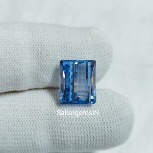 Puede incluir: Una gema rectangular de color azul claro con una superficie facetada. La gema se muestra sobre una tela blanca. El texto "SahergemsIN" es visible en la tela debajo de la gema.