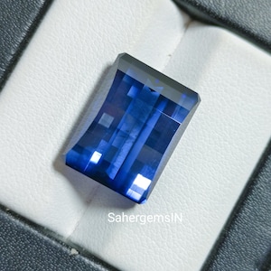 May include: A rectangular, faceted sapphire gemstone in a deep blue shade. The gem is set on a white, textured surface within a black box. The text "SahergemsIN" is visible on the surface.