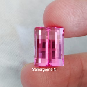 May include: A rectangular, faceted pink gemstone held between fingers. The gem has a clear, crystalline appearance with internal reflections. The text "SahergemsIN" is visible. The gem is a vibrant shade of pink.