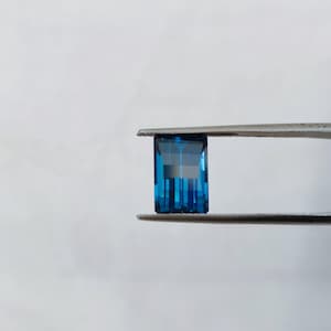 Puede incluir: Una piedra preciosa azul de forma rectangular, sostenida por unas pinzas sobre un fondo blanco.