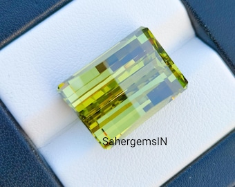 Può includere: Una pietra preziosa verde e gialla di forma rettangolare, esposta in una scatola nera e bianca. La pietra preziosa ha un taglio sfaccettato e una finitura lucida. Il testo "SahergemsIN" è visibile nell'angolo in basso a destra dell'immagine.