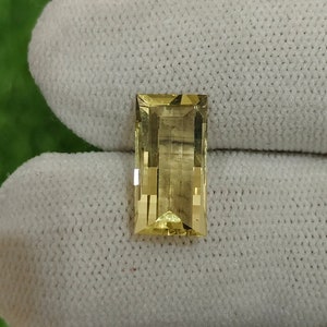 May include: A rectangular, light yellow gemstone with a faceted cut. The gemstone is displayed on a white fabric background.