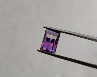 Può includere: Una pietra preziosa viola di forma rettangolare è tenuta da una pinzetta. La pietra preziosa ha un leggero luccichio ed è traslucida.