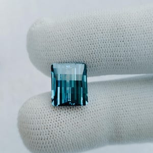 Op de afbeelding: Een vierkante, gefacetteerde edelsteen met een blauwgroene tint. De edelsteen is geplaatst tegen een witte achtergrond.