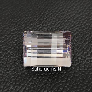 May include: A clear, faceted, rectangular gemstone with a light purple hue. The gemstone is set against a black background.
