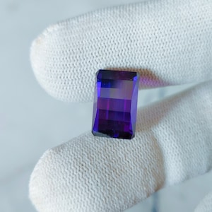 Könnte beinhalten: Ein tiefvioletter Edelstein mit rechteckiger Form, in Facetten geschliffen. Der Edelstein wird auf einem weißen Stoffhintergrund präsentiert.