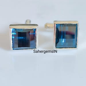 May include: A pair of square cufflinks with a silver-colored frame and a deep blue, faceted center. The cufflinks are displayed on a white surface. The text "SahergemsIN" is visible.
