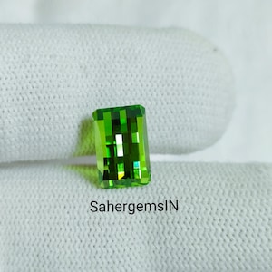 May include: A rectangular, faceted, bright green gemstone. The gem is set against a white, textured background. The text "SahergemsIN" is printed in black below the gemstone.