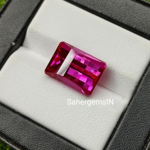 Op de afbeelding: Een levendige roze edelsteen met een rechthoekige snede, gepresenteerd in een witte fluwelen doos. De doos is bekleed met zwart leer en heeft de tekst "SahergemsIN" gedrukt op de bodem.