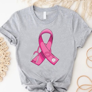 May include: A gray t-shirt with a pink ribbon and a stethoscope design. The ribbon is a symbol of breast cancer awareness.