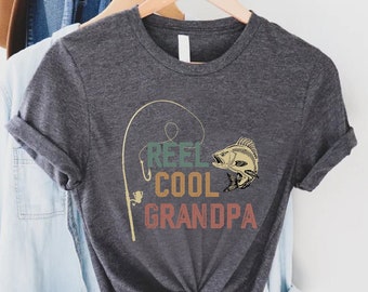 May include: A dark gray t-shirt with a vintage-style graphic of a fishing rod and a fish. The text "Reel Cool Grandpa" is printed on the shirt.