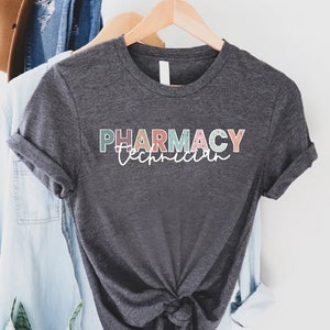 May include: A dark grey t-shirt with the text "PHARMACY Technician" in colorful letters. The letters are outlined in white and have a speckled texture.