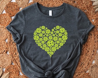 Puede incluir: Una camiseta gris oscuro con un diseño en forma de corazón hecho de bolas de pickleball verdes y amarillas.