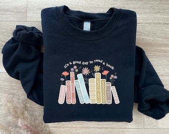 Puede incluir: Una sudadera negra con un gráfico de una pila de libros coloridos con flores y el texto "Es un buen día para leer un libro".
