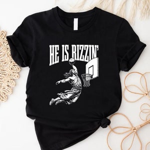 May include: Black t-shirt with a white graphic of a person dunking a basketball. The text "HE IS RIZZIN'" is above the graphic.