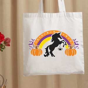 Op de afbeelding: Witte tote bag met een zwart silhouet van een eenhoorn die op een regenboog staat met vleermuizen en pompoenen. De eenhoorn heeft een spin op zijn hoorn.