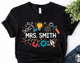 May include: Black t-shirt with a science theme design. The design includes a lightbulb, DNA strand, rocket, and other science-related icons. The text "MRS. SMITH" is printed on the shirt.
