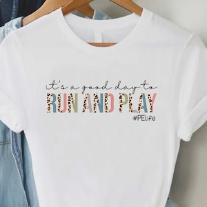 May include: White t-shirt with the text "it's a good day to RUN AND PLAY #PELife" printed on the front. The text is in a colorful font with a leopard print pattern.